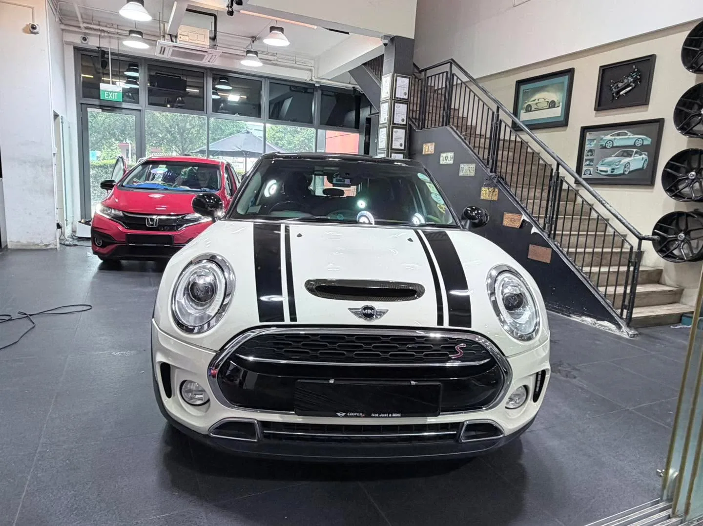 A white Mini hatchback with black racing stripes and a red Honda sedan parked in the workshop.