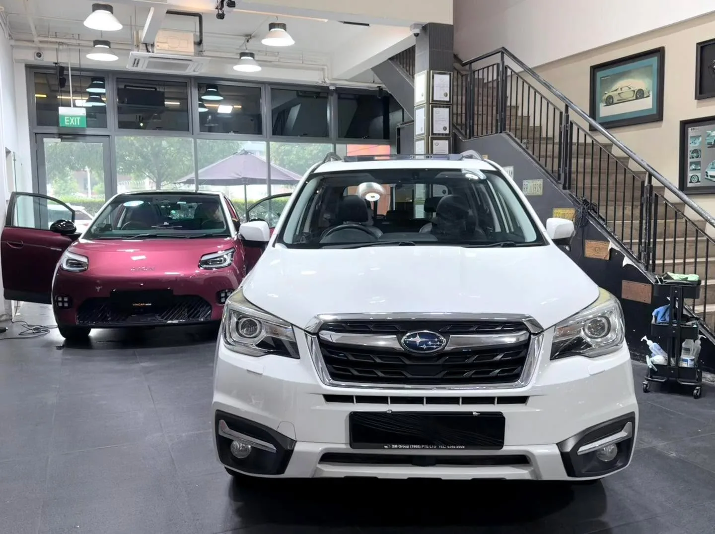 A white Subaru Forester viewed front-on in the workshop, with a maroon hatchback visible behind it.