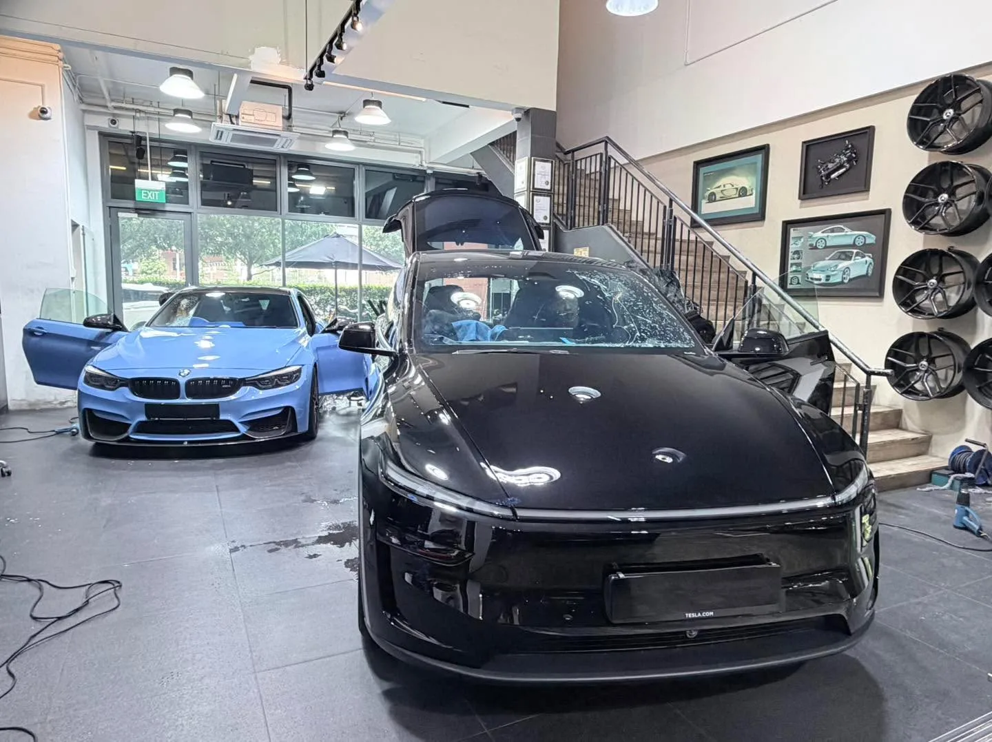 A black Tesla with a blue M performance BMW to the left, parked in the workshop.