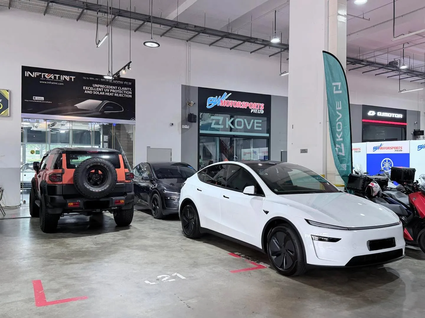 An orange and black SUV, a dark saloon, and a white Tesla parked in a workshop space.