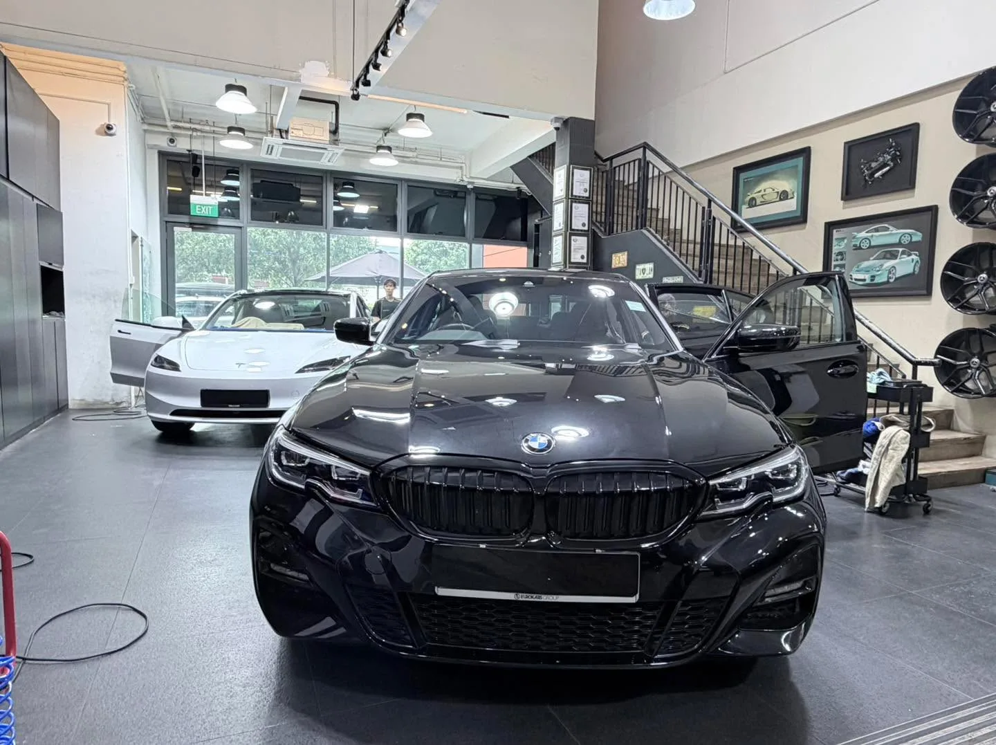 A black BMW saloon facing forward in a workshop with a white Tesla behind it.