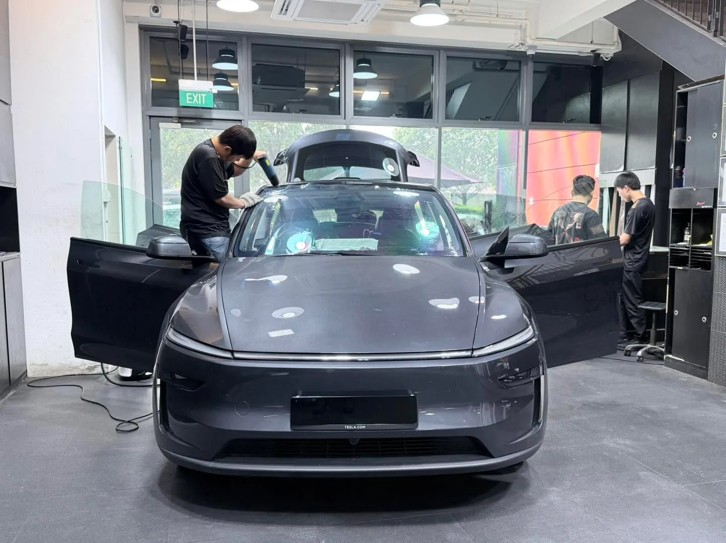 A dark grey Tesla with doors open and technicians applying film to the windows.