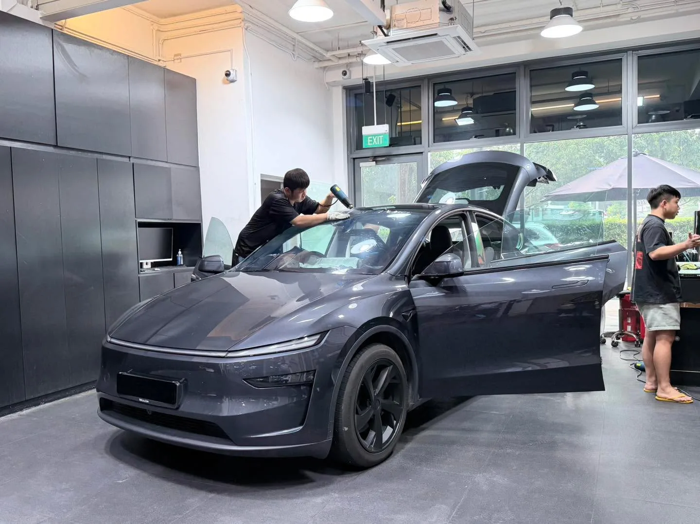 A dark grey Tesla with open doors and technicians working on the window tinting.