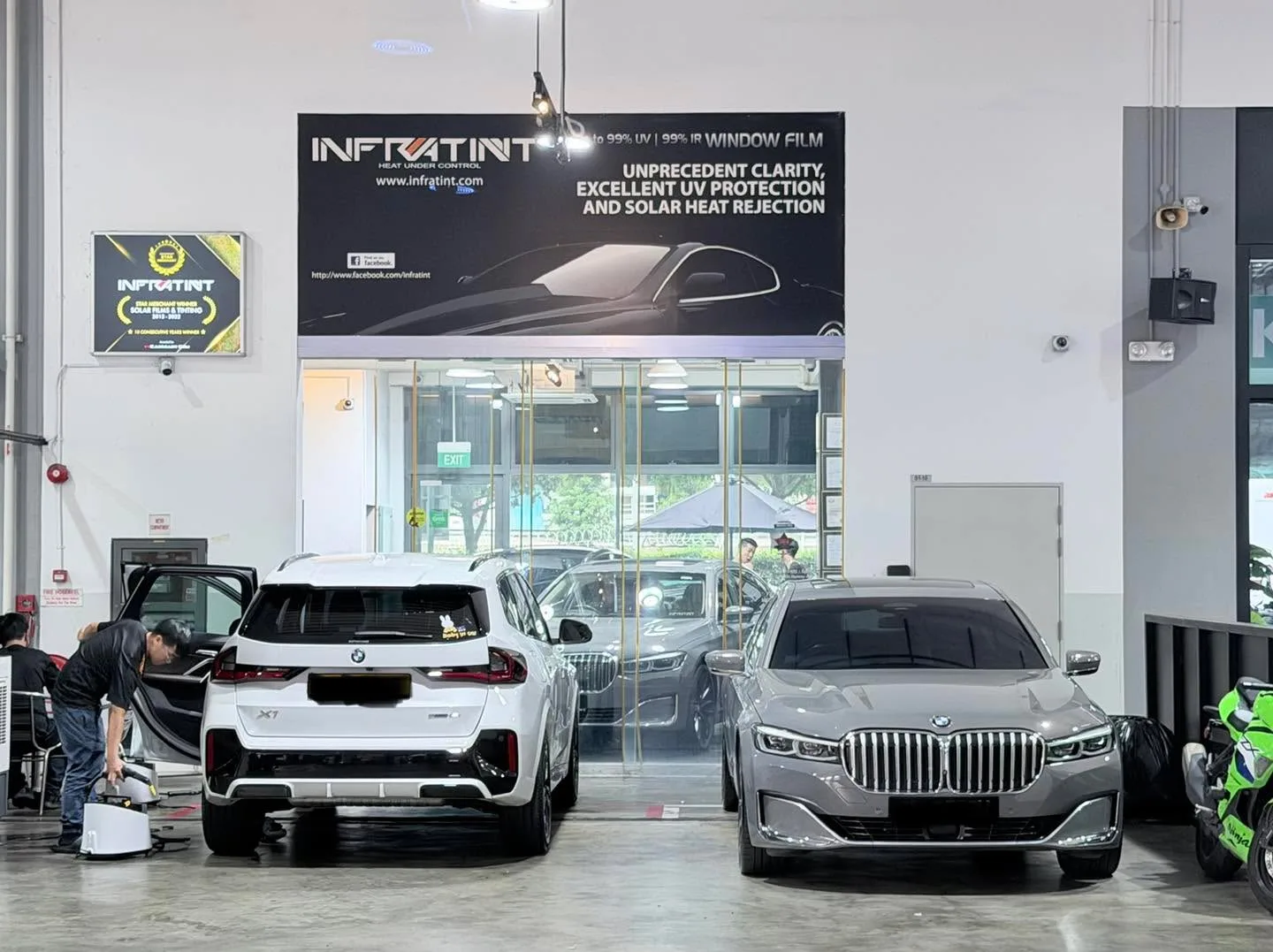 A white BMW X1 SUV and a grey BMW sedan being prepared inside the workshop.