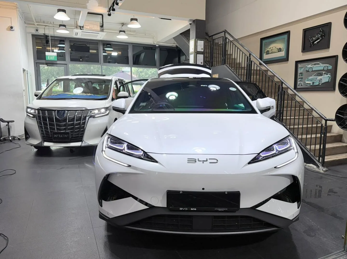 A white BYD sedan and a white Toyota Alphard multi-purpose vehicle in an indoor facility with artwork on walls.