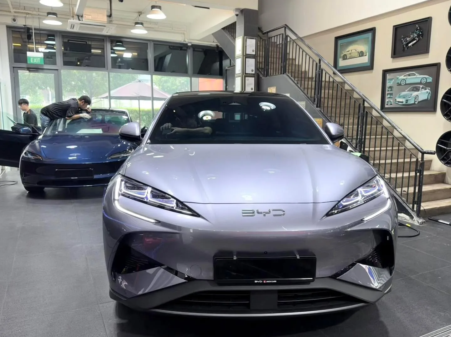 A silver BYD sedan and a white Tesla sedan in the facility with people working nearby.