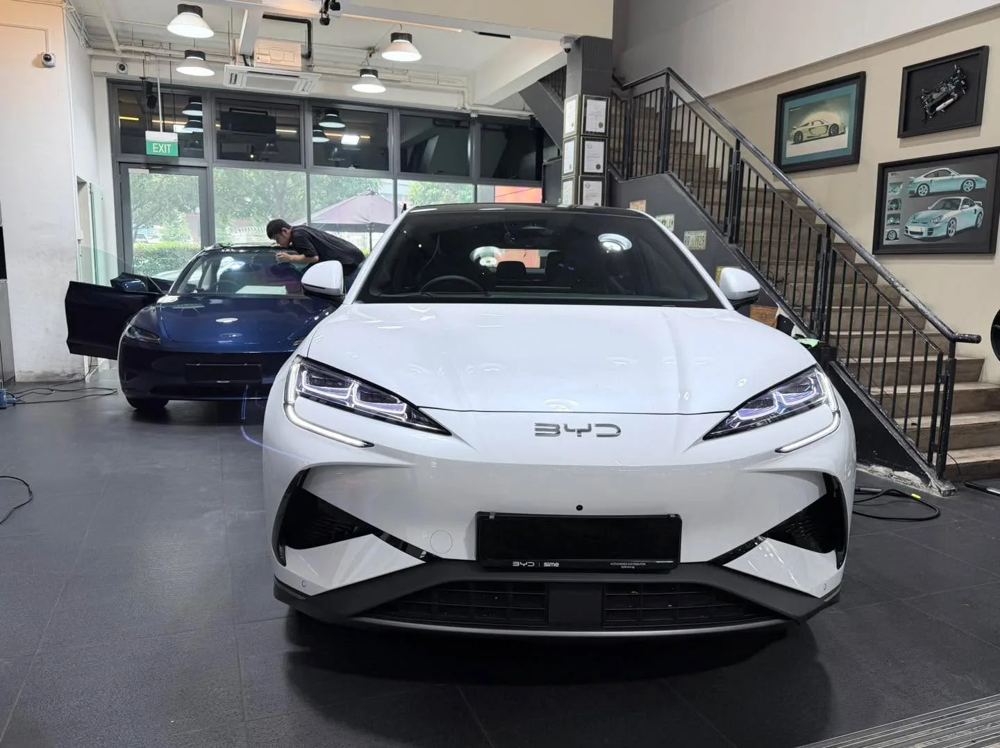 A white Tesla saloon in the foreground with an MG hatchback visible behind in the workshop.
