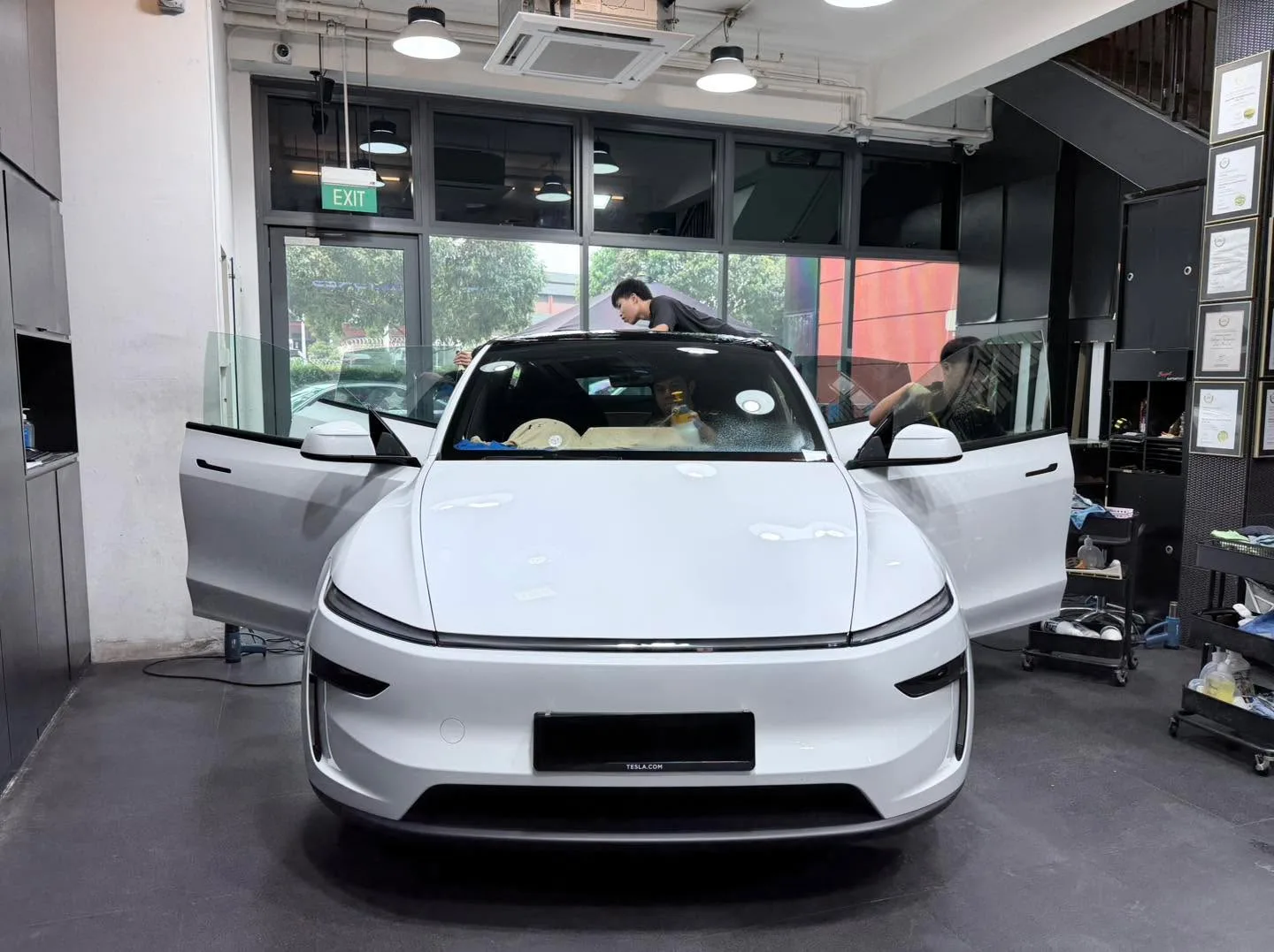A white Tesla hatchback with open doors sits in the workshop with technicians working on its windows.