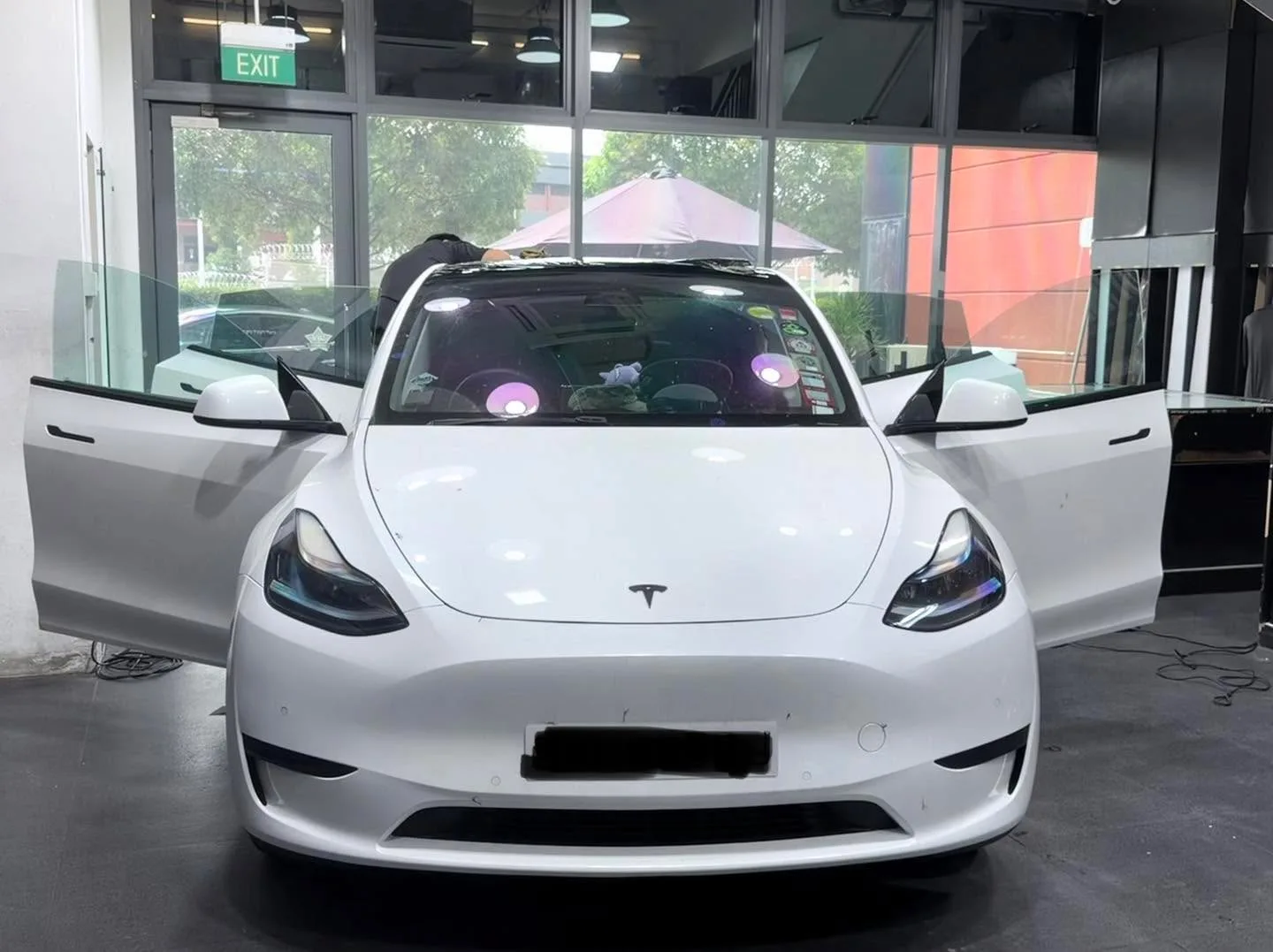 A white compact sedan with gullwing-style doors open, viewed from the front in the workshop.