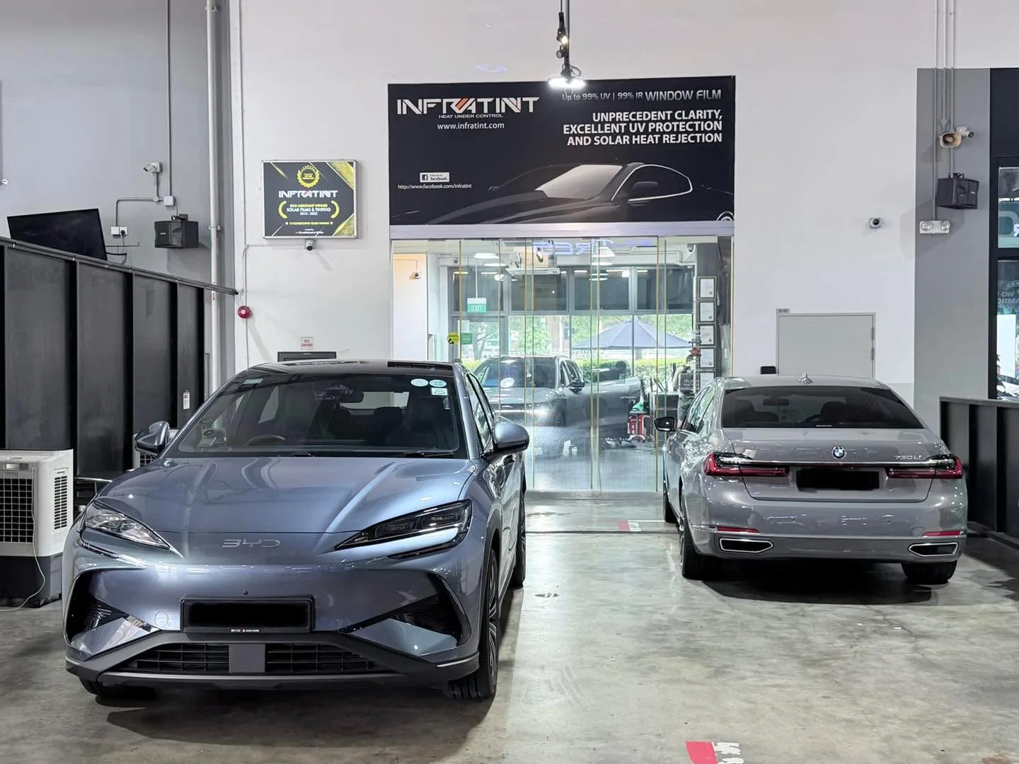 A silver sedan and a grey sedan in the workshop bay with glass doors and Infratint signage behind.