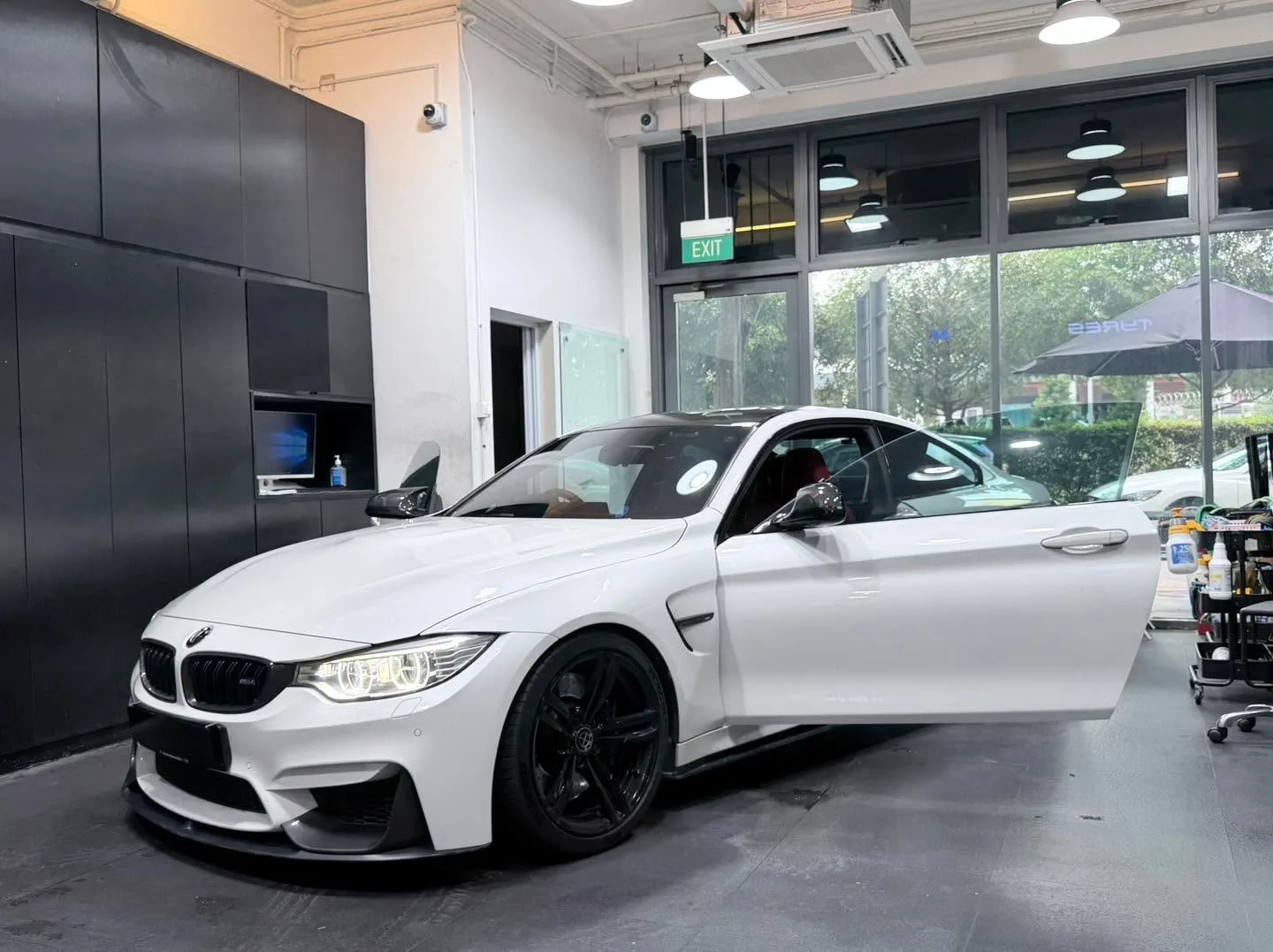 A white compact coupe with open door and workshop equipment nearby.