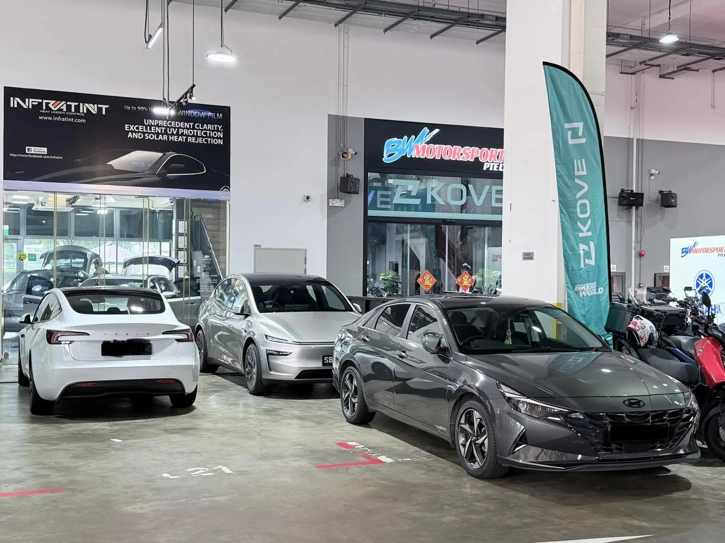 Three cars parked in a workshop. From left: white Tesla Model Y, silver Tesla Model 3 Highland, and dark grey Hyundai Avante.