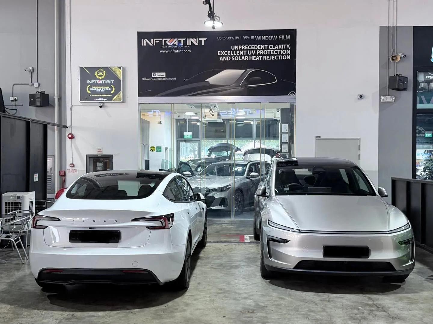 Front view of two cars parked indoors. Left: white Tesla Model Y. Right: silver car with distinctive grille.