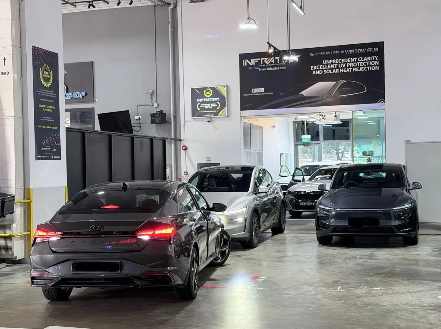 Three cars parked in a workshop. Left: dark red Hyundai Avante with lights on. Centre: silver car. Right: dark vehicle.