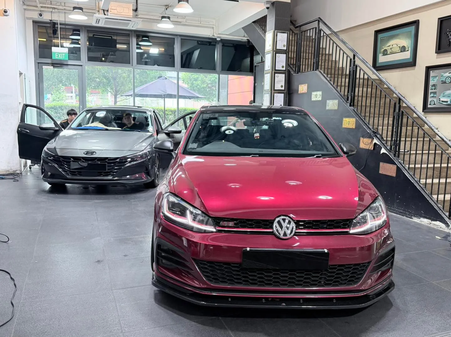 Two cars parked indoors. Left: grey Hyundai Avante with door open. Right: red Volkswagen Golf GTI.