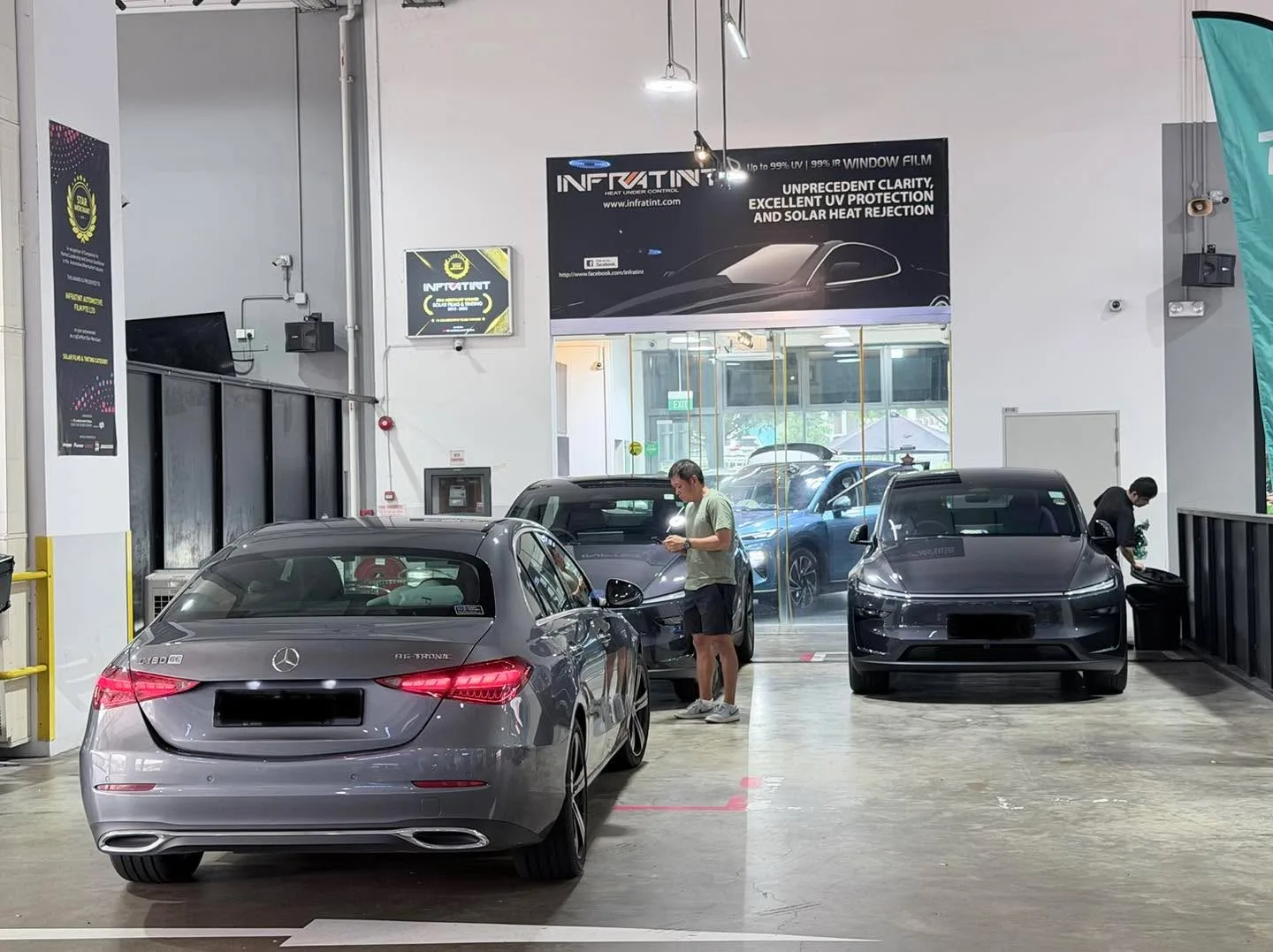 A grey Mercedes-Benz sedan with staff working on its windows, and a dark sports car beside it.