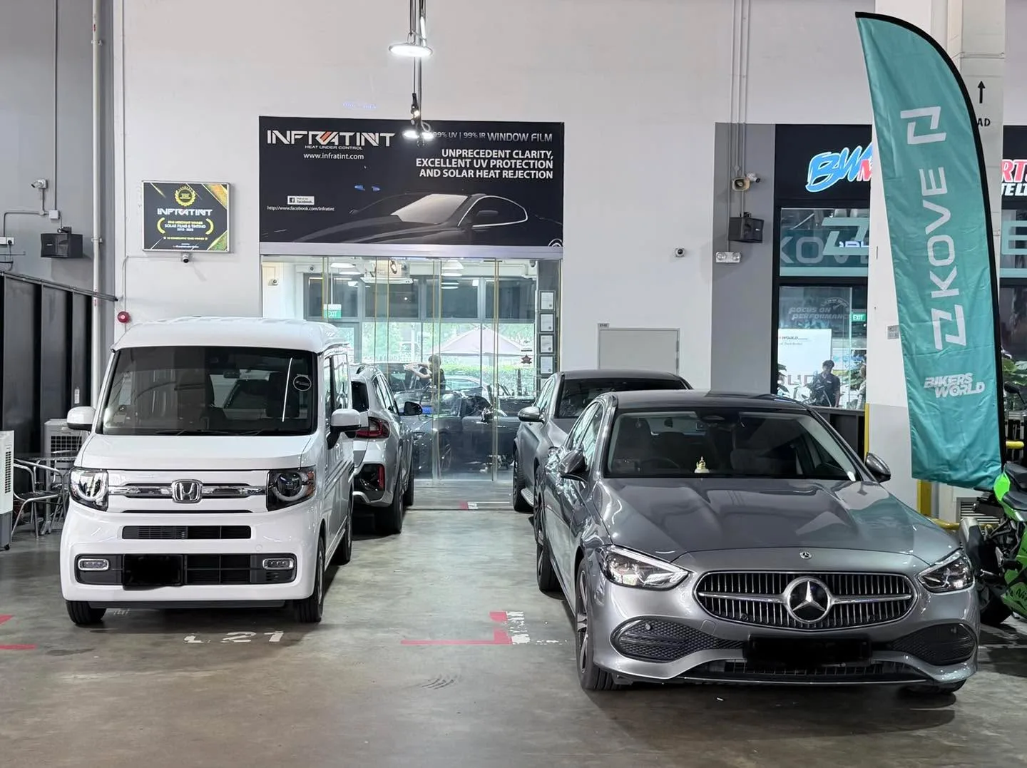 A white Honda compact vehicle on the left and a silver Mercedes-Benz sedan on the right in the workshop.