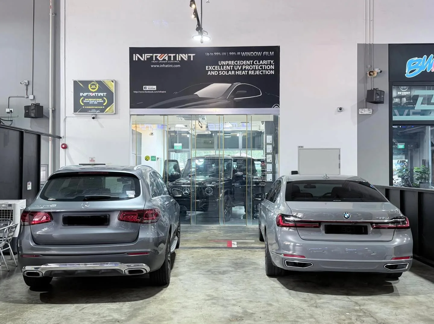 A black Mercedes-Benz G63 SUV and a silver BMW sedan parked in the workshop exterior bay from a different angle.