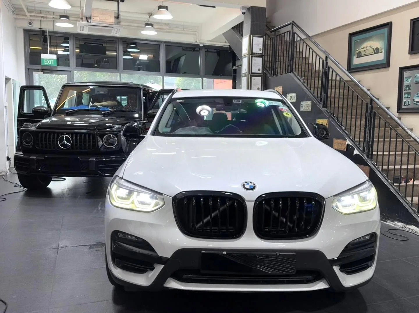 A white BMW X3 SUV viewed from the front in the workshop interior, with a black Mercedes-Benz vehicle visible behind it.
