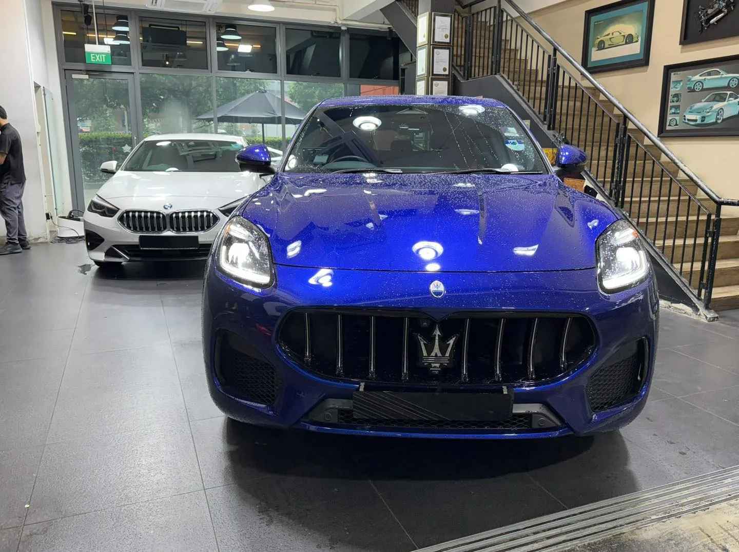 A blue Maserati saloon with distinctive grille, parked indoors with a white BMW sedan visible behind.