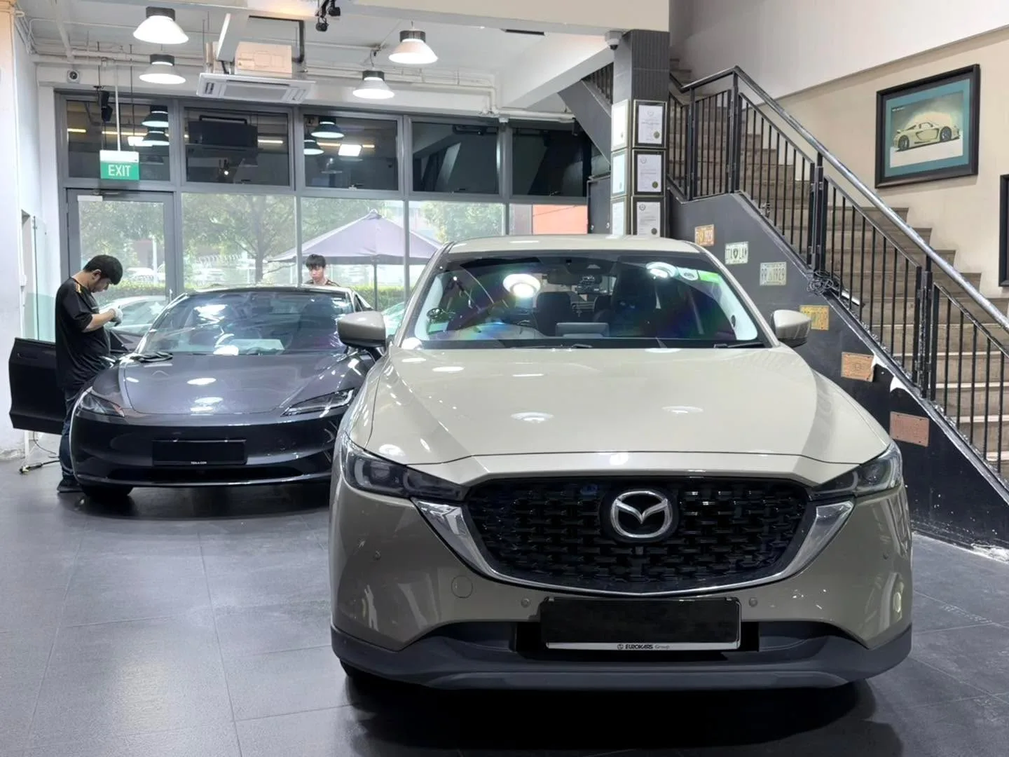 A silver Mazda CX-5 and a black car parked in the showroom with staff working nearby.