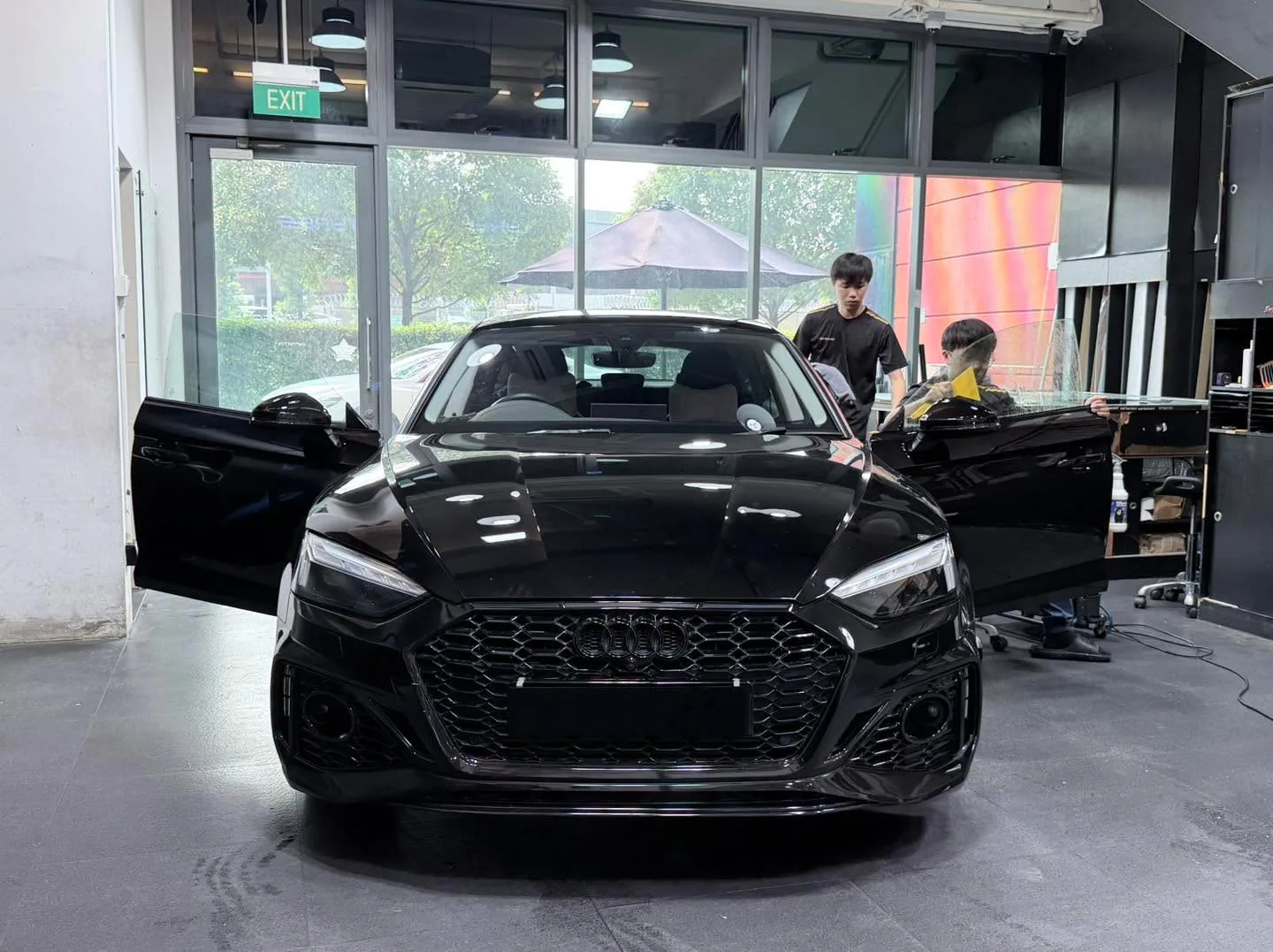 A black Audi A5 viewed from the front in the workshop, with open doors and staff nearby.