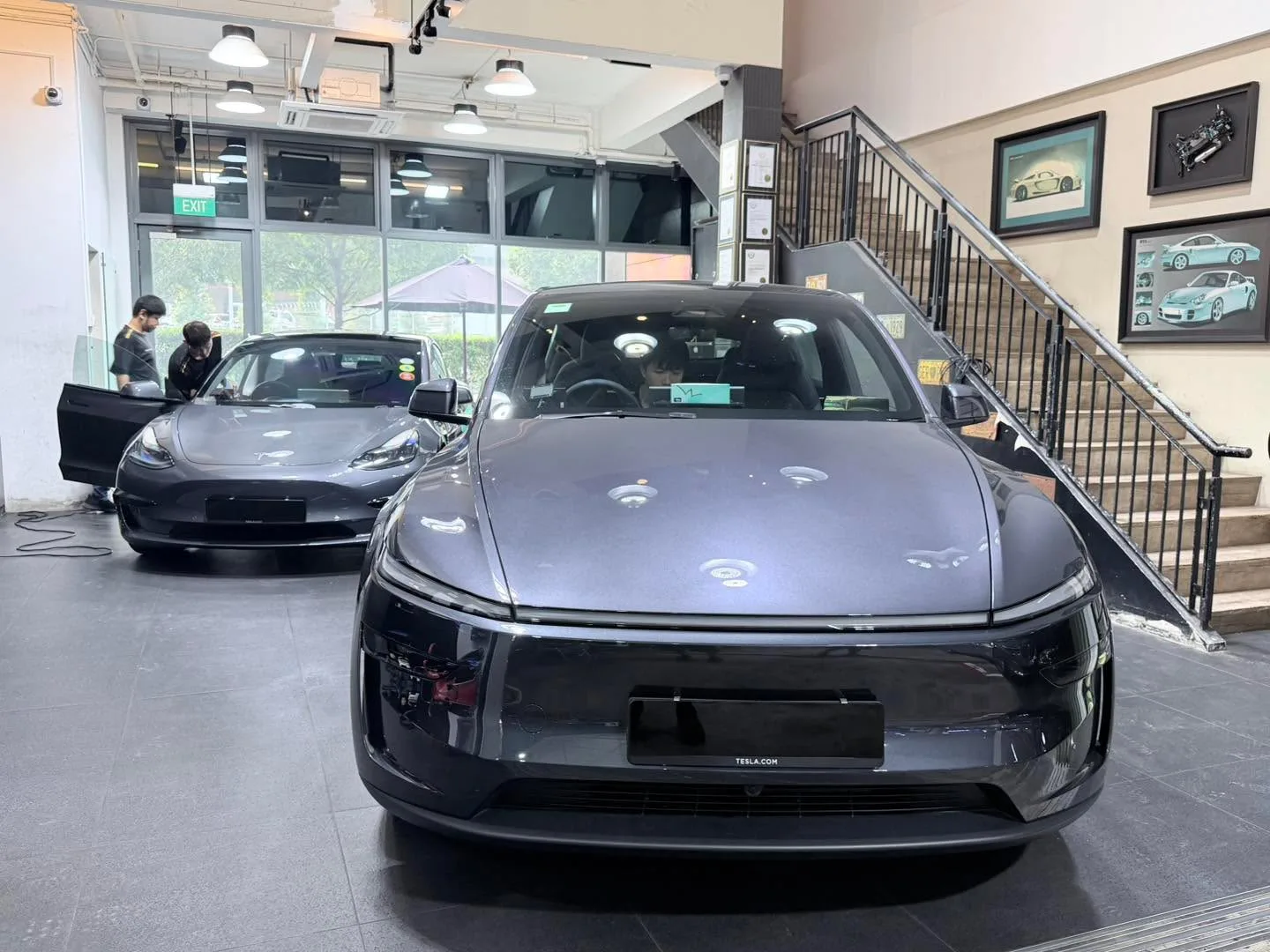 A grey Tesla Model Y facing forward in the workshop, with a dark-coloured Tesla Model 3 to the left.