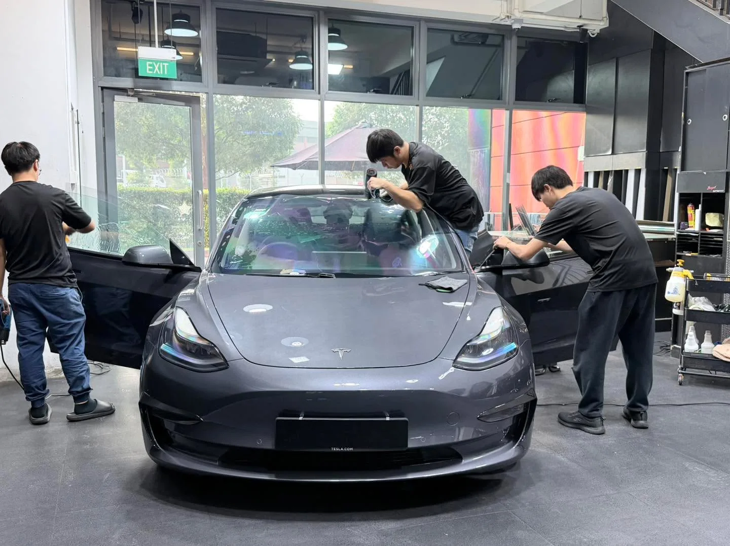 A dark-coloured Tesla with technicians working on the windows, doors open.