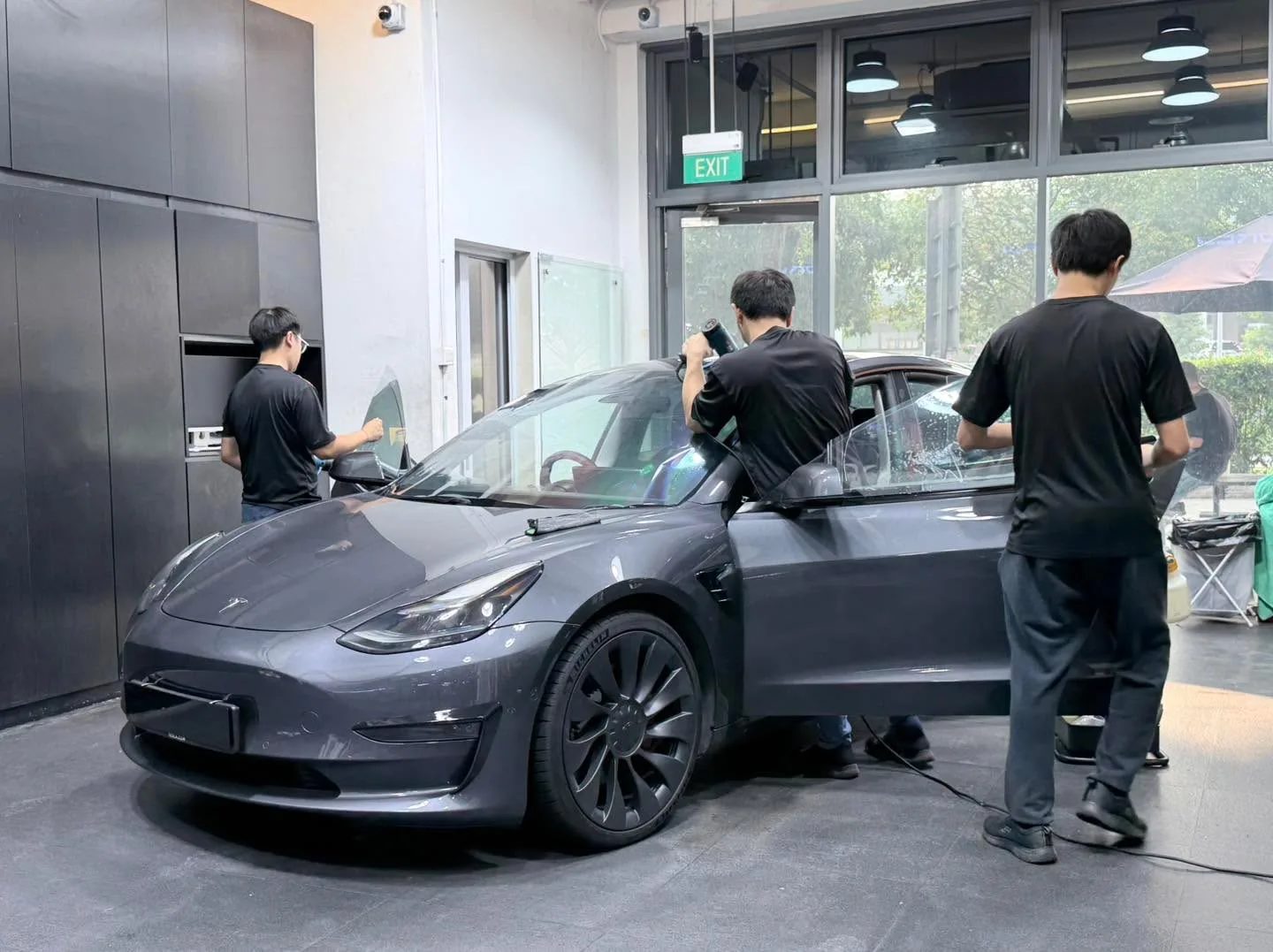 A dark grey Tesla sports car with technicians applying window tint to the doors and windows.
