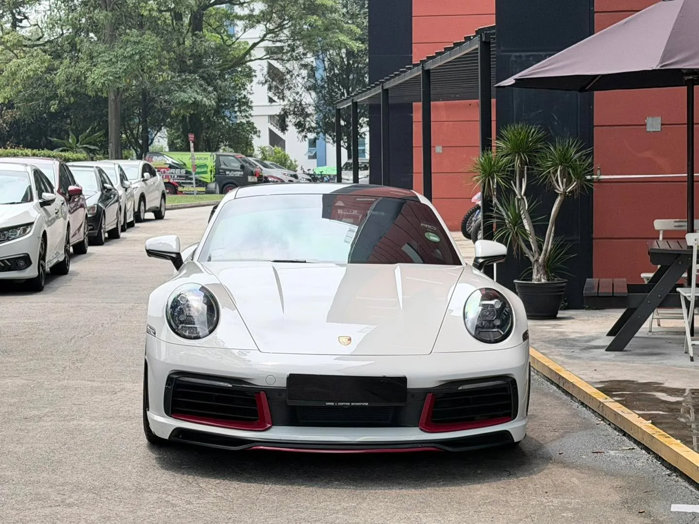 A white Porsche 911 with red accent details parked in the workshop carpark, with other white vehicles visible in the background.