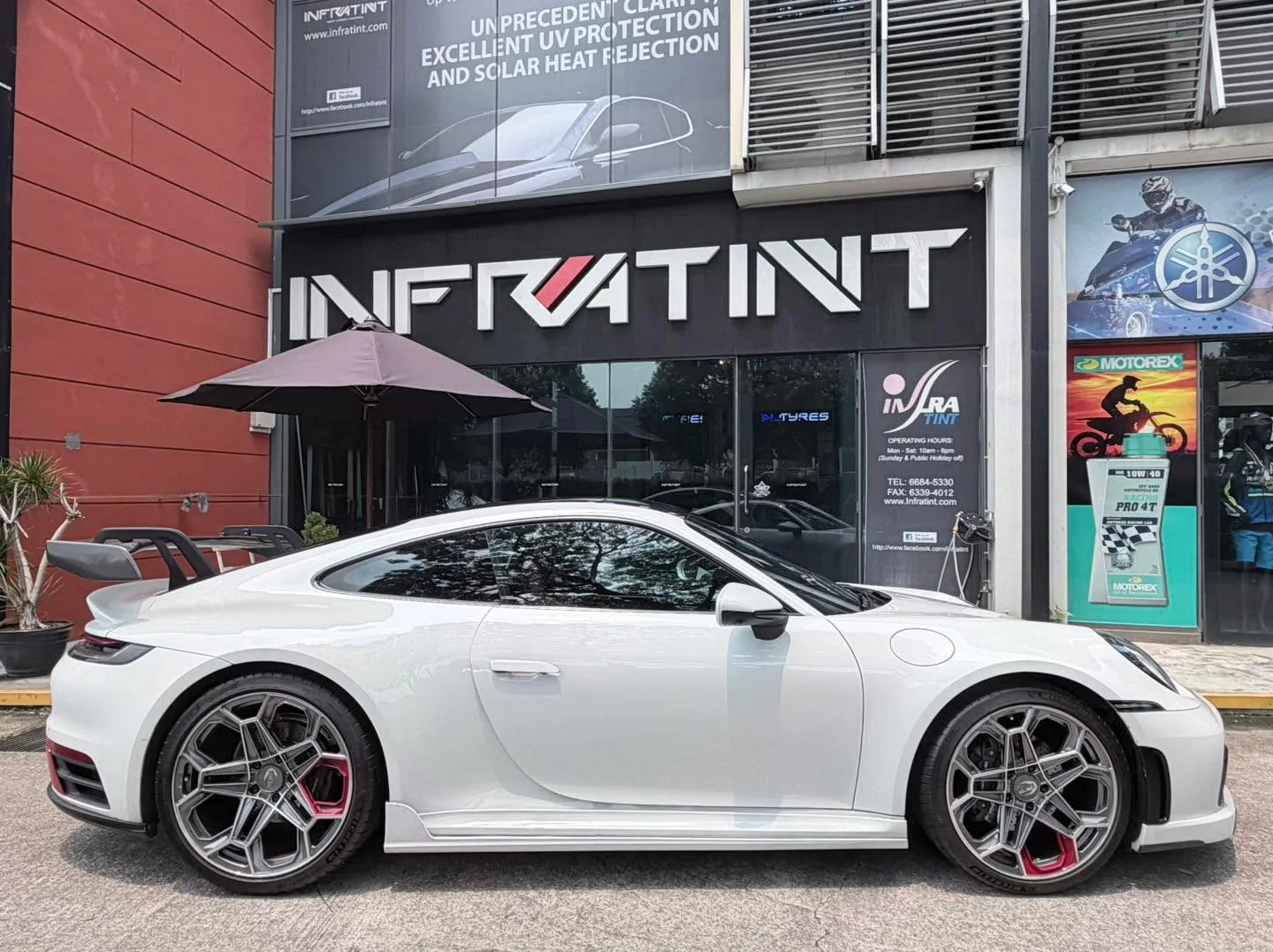 The white Porsche 911 parked outside the Infratint workshop storefront, viewed from the side.