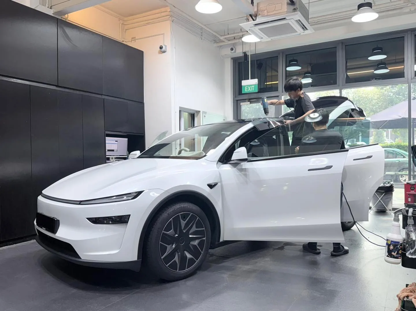 A white Tesla sports car with an open door, undergoing window tinting as a technician applies film to the side windows.