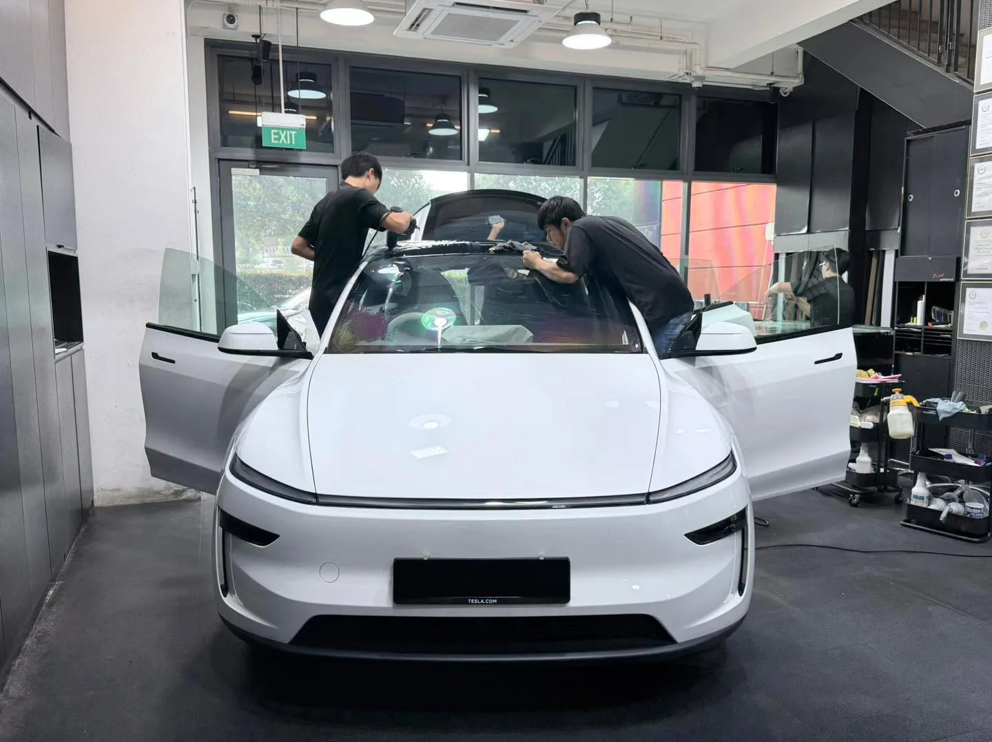 A white Tesla sedan with its doors open, being worked on by two technicians applying window tint inside the workshop.