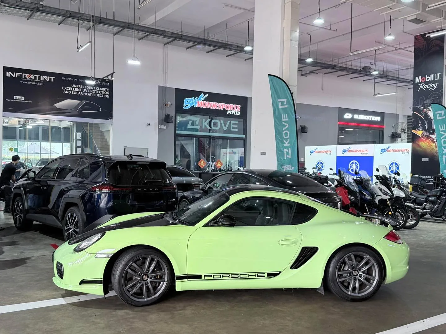 A lime-green Porsche Cayman coupe and dark blue SUV at a workshop with exposed ceiling and signage.