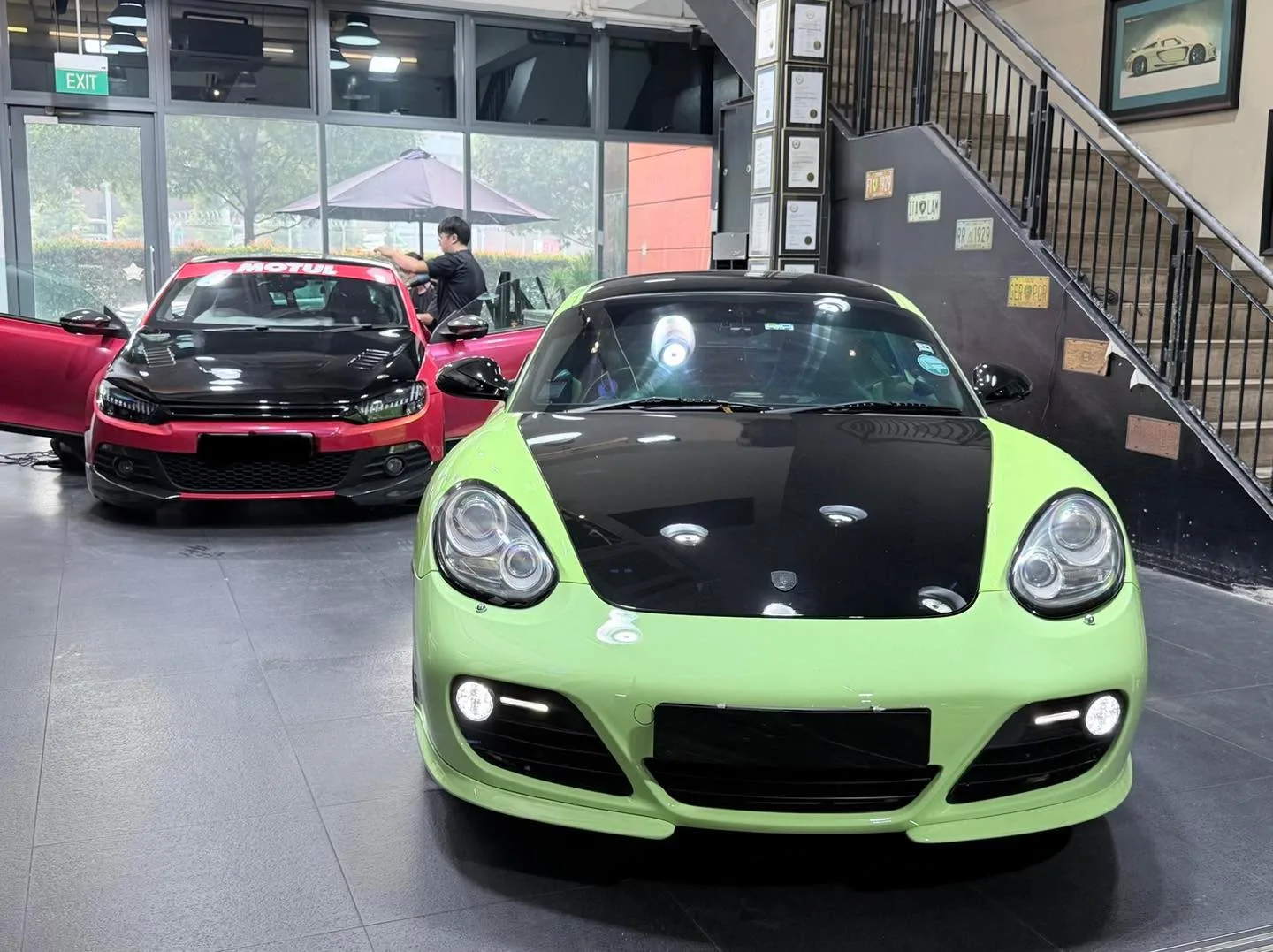 Lime-green Porsche Cayman with black hood and red sedan visible behind it in workshop.