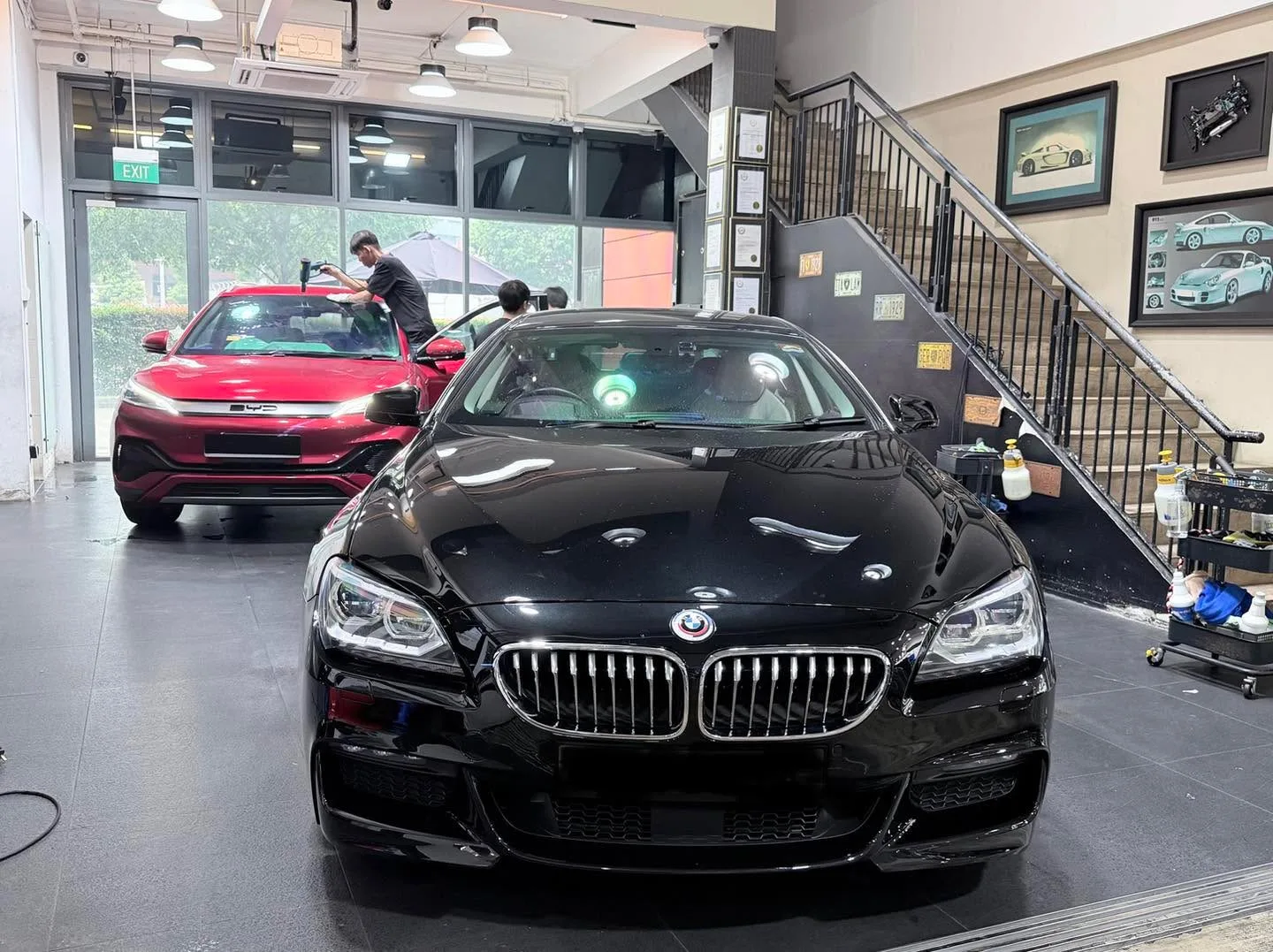 Black BMW 6 Series sedan and burgundy car with staff working nearby in workshop.