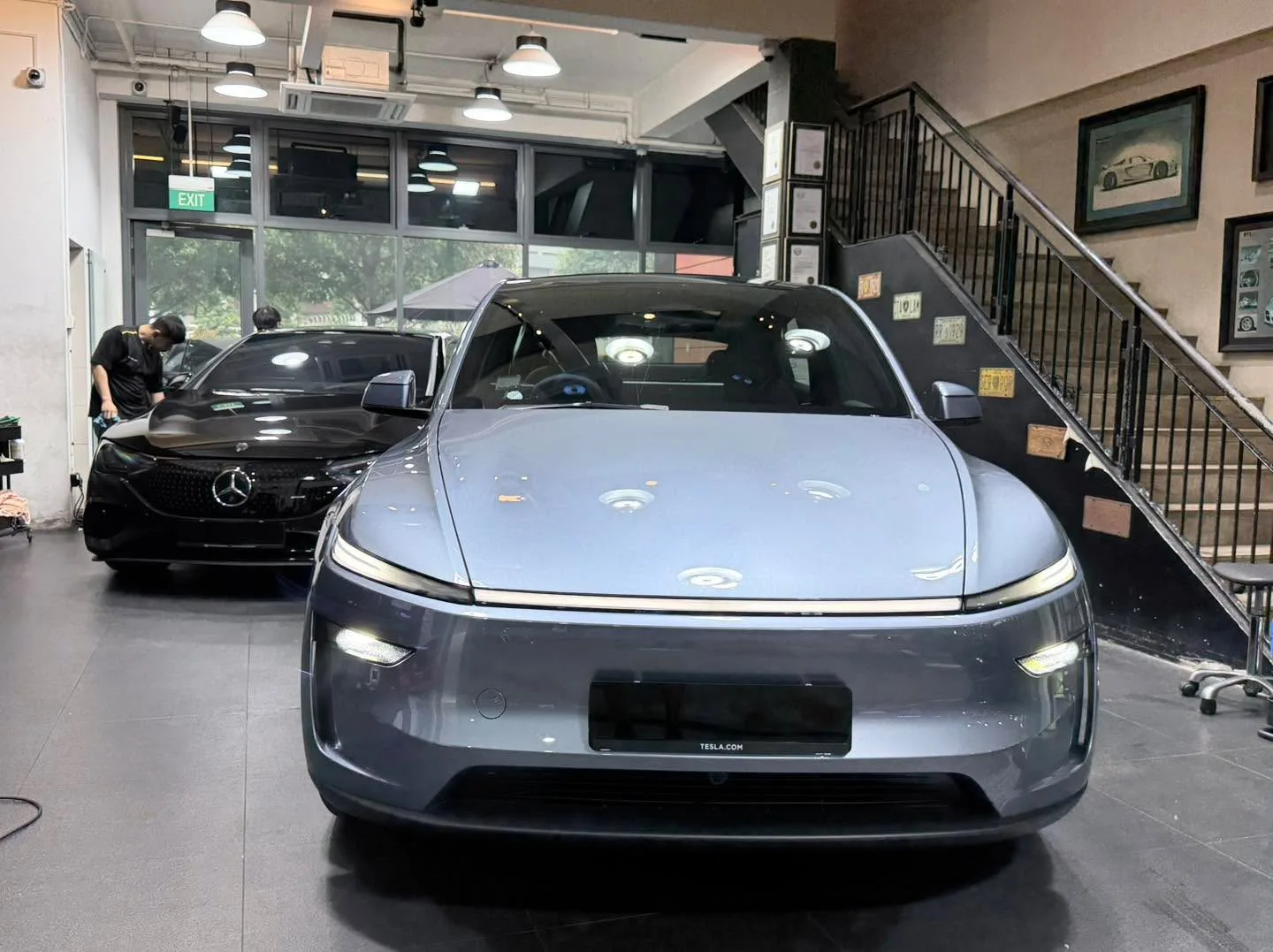 A light silver Tesla sedan in the foreground and a dark Mercedes-Benz sedan behind it in the workshop.