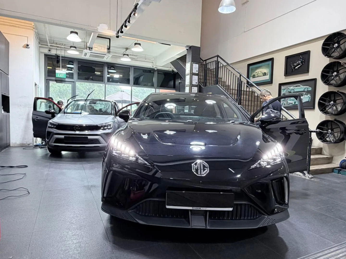 A black MG sedan with doors open in the foreground, and a light-coloured car behind it in the workshop.