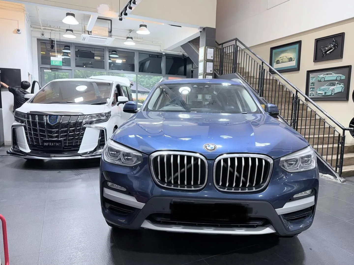 A blue BMW X3 in the foreground facing the camera, with a white Toyota Alphard visible behind.
