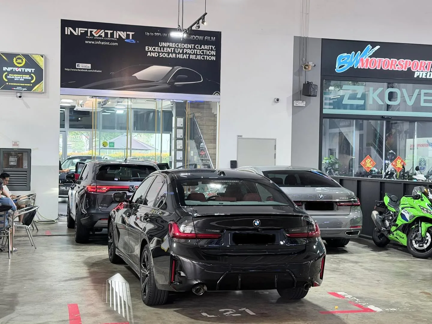 A black BMW saloon and a grey car in an open workshop with glass frontage and Infratint signage.