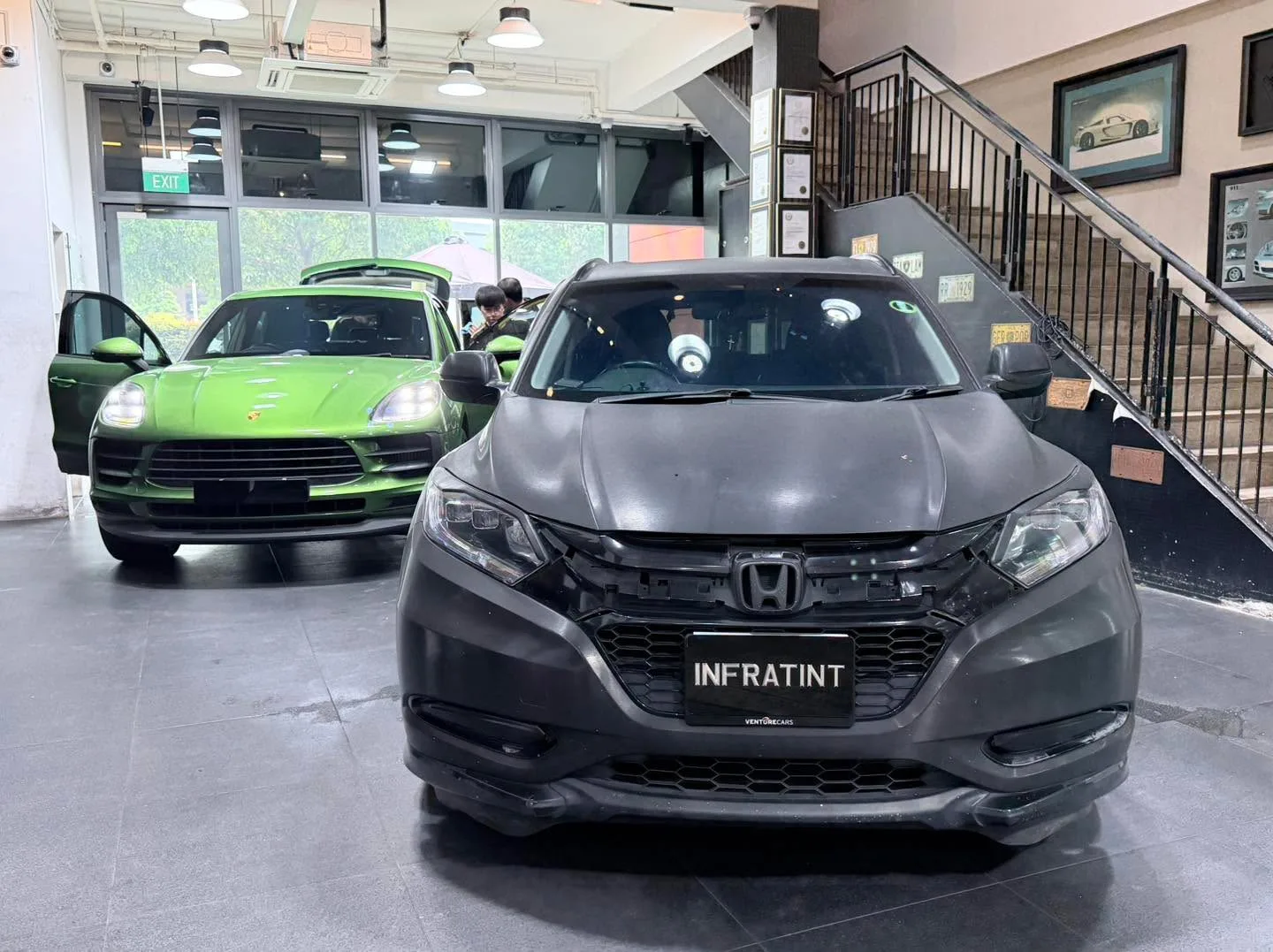 A green Porsche Macan S with doors open, a dark grey Honda Vezel ahead, and staff working inside.
