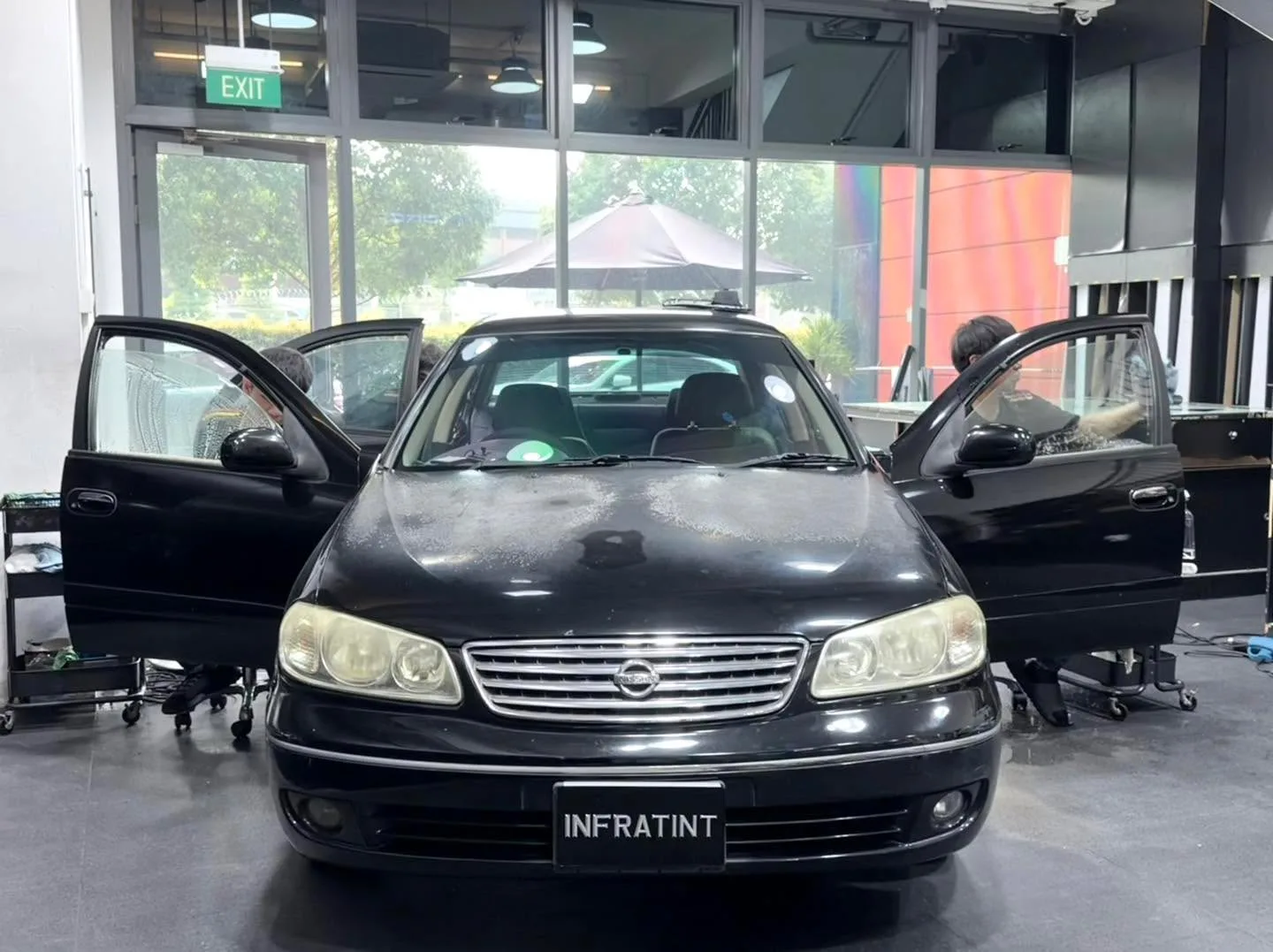 A black Nissan Sunny sedan with driver side door open in the workshop.