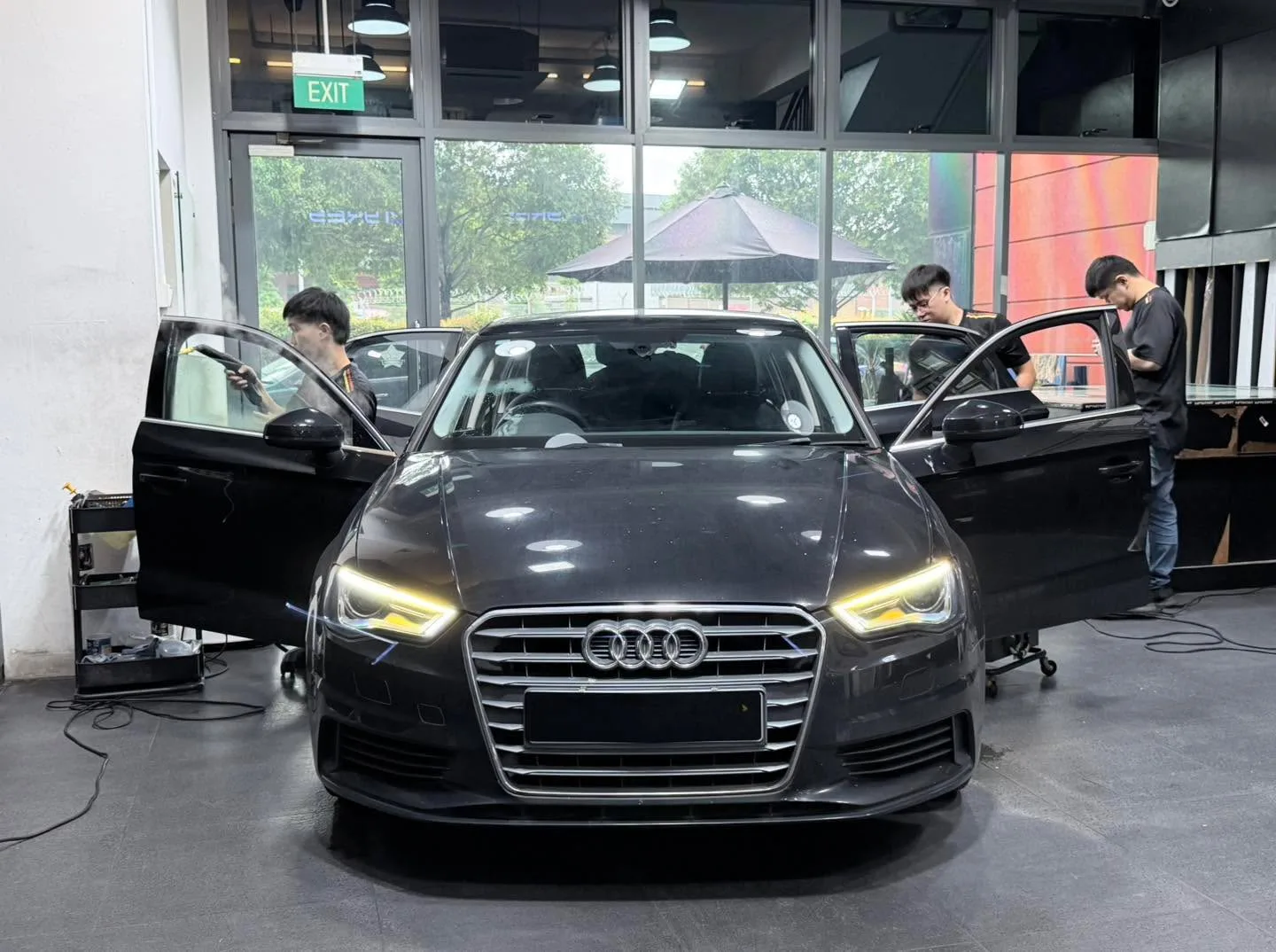 A dark grey Audi sedan with doors open, technicians working on windows.