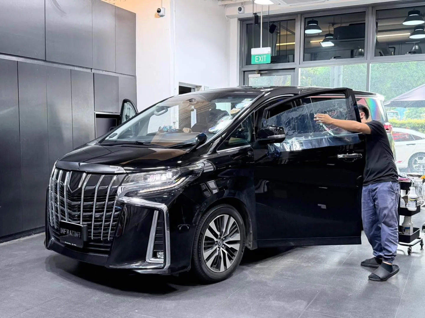 A black Toyota Alphard with technicians applying film to the windows.