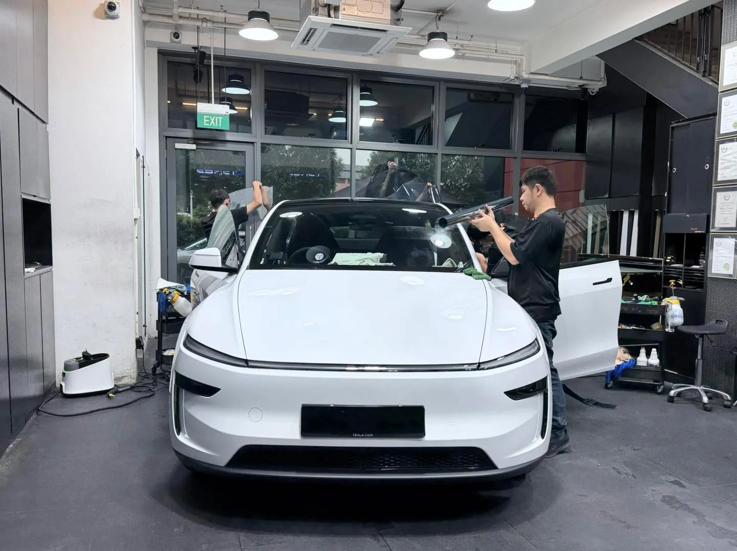 A white Tesla Model Y with doors open and technicians working on windows.
