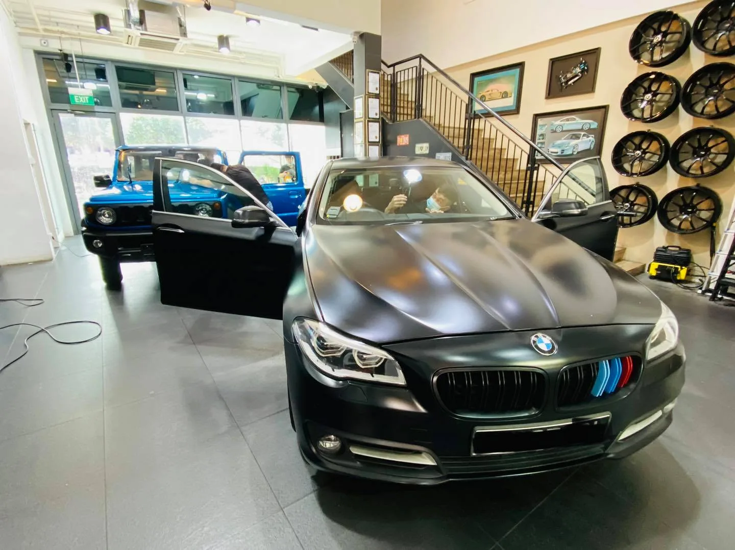 Matte black BMW sedan and blue Suzuki Jimny inside Infratint Singapore beside the wheel display