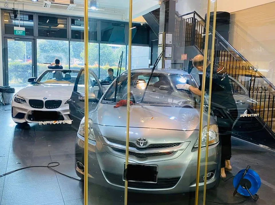 Technician tinting a silver Toyota Vios with a white BMW 2 Series behind at Infratint Singapore